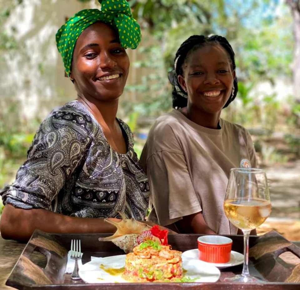To damer i Afrika sitter foran et brett med mat og drikker, mens de smiler til kameraet.