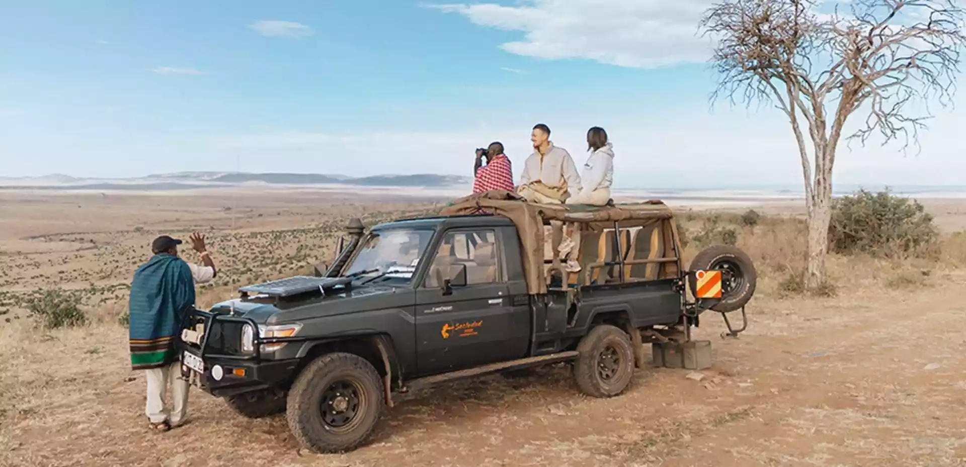 Reisende som speider utover savannen på baksiden av en safari jeep, med guide som speider foran bilen.