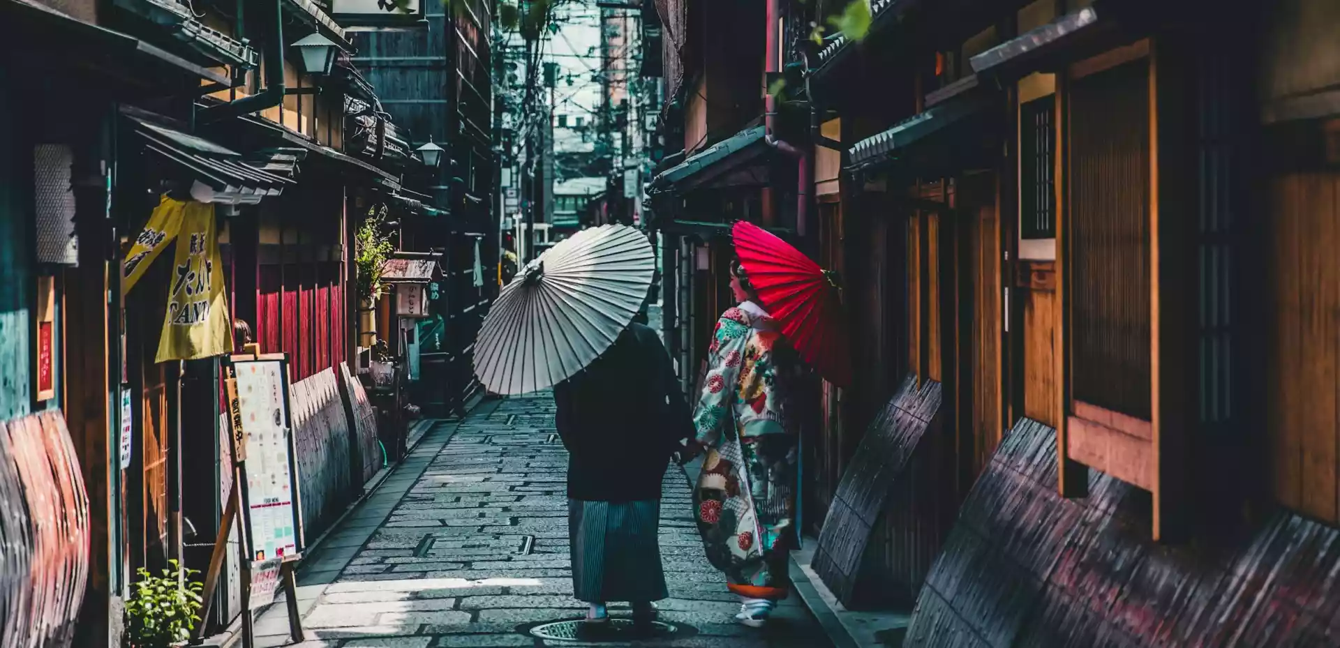 En brolagt bakgate i Kyoto, omgitt av små japanske hus. To damer går fra kamera, begge bærer hver sin papir paraply.