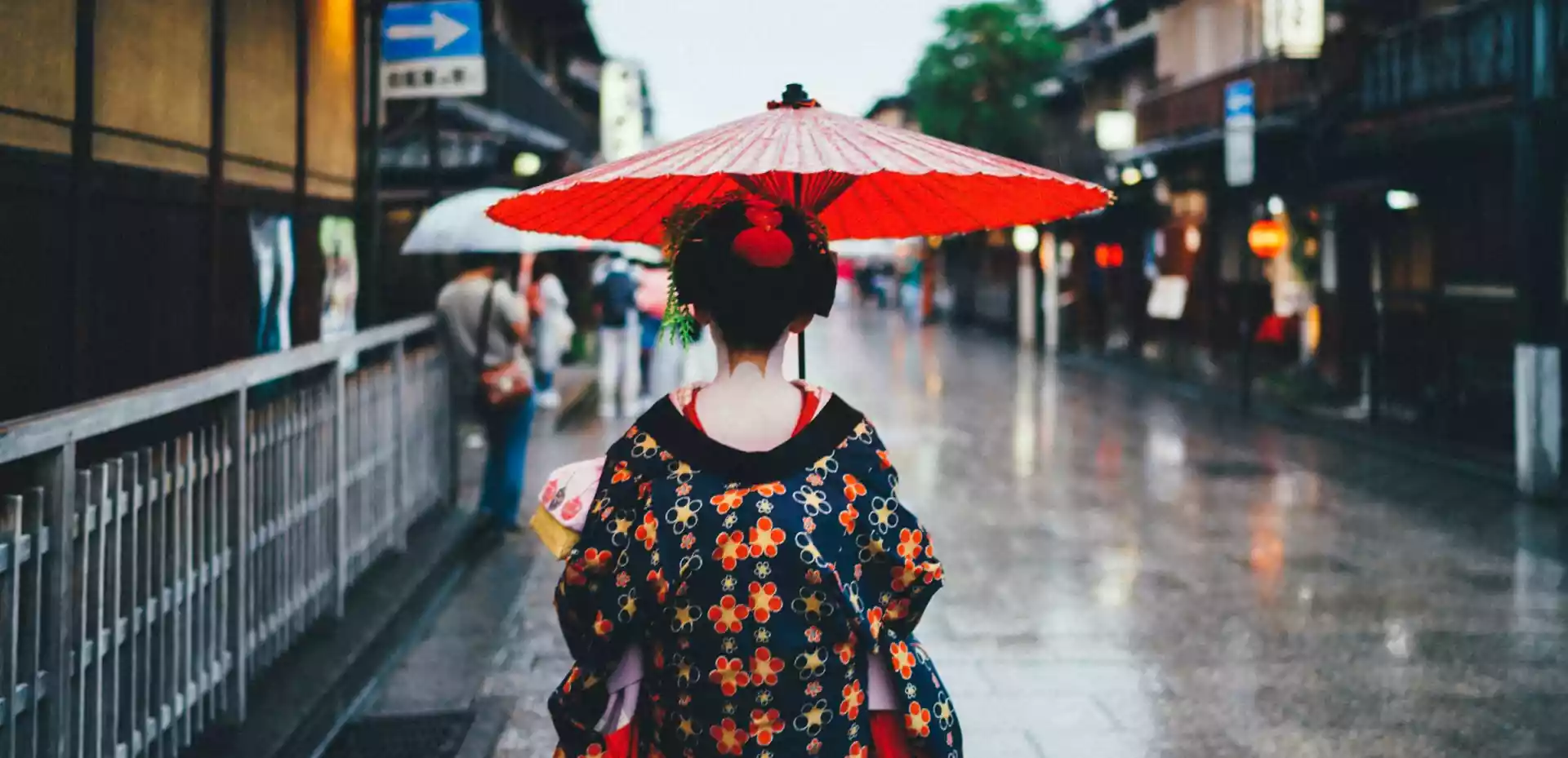 En kvinne med en rød paraply går gjennom en japansk gate i regnværet. Hun står med ryggen til kameraet, og paraplyen skaper et levende fokus mot det omkringliggende landskapet.