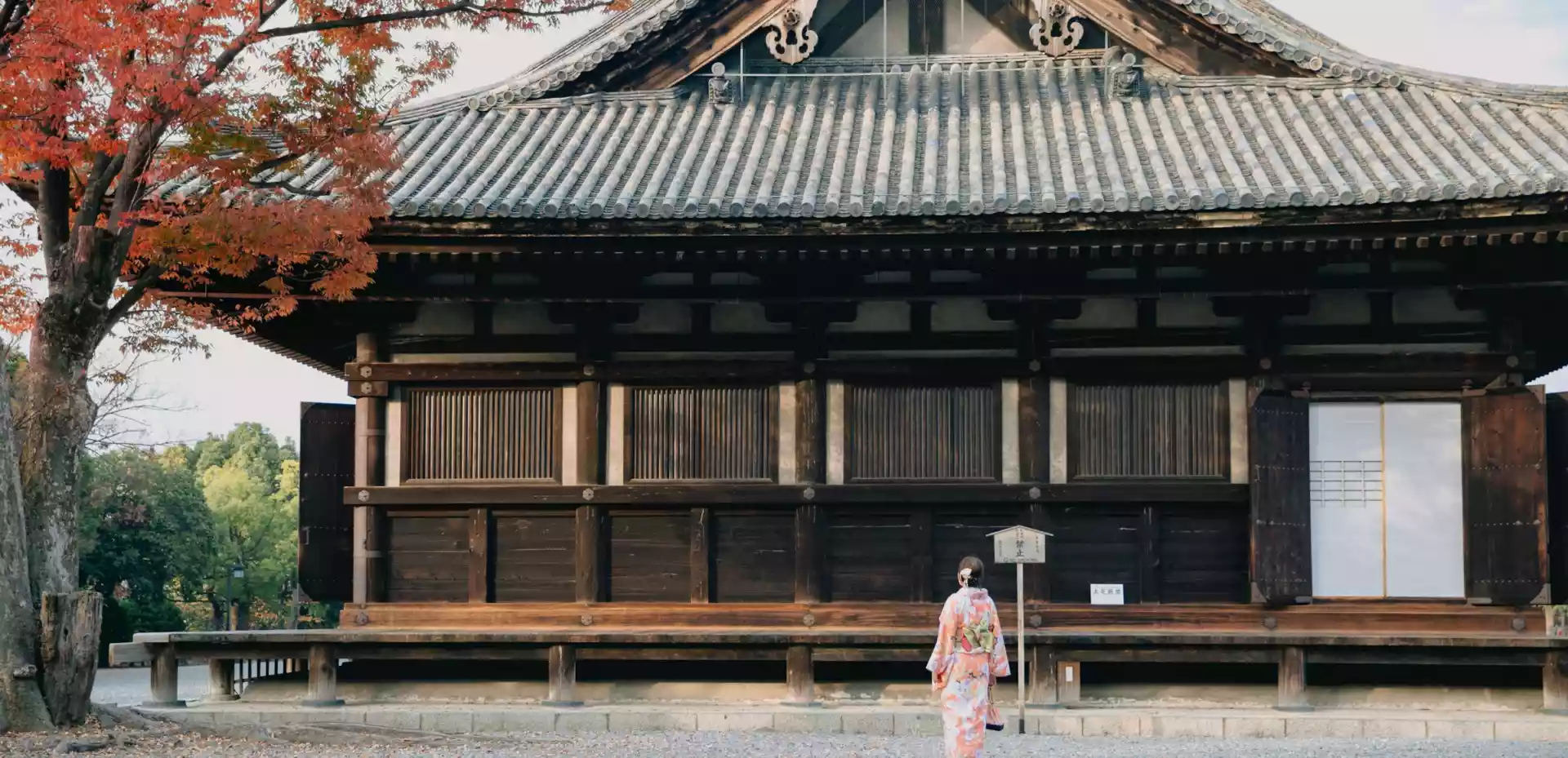 Et hus i Kyoto i tradisjonell japansk stil. På den åpne plassen foran huset står en kvinne med rygge til, kledd i tradisjonell japansk drakt.
