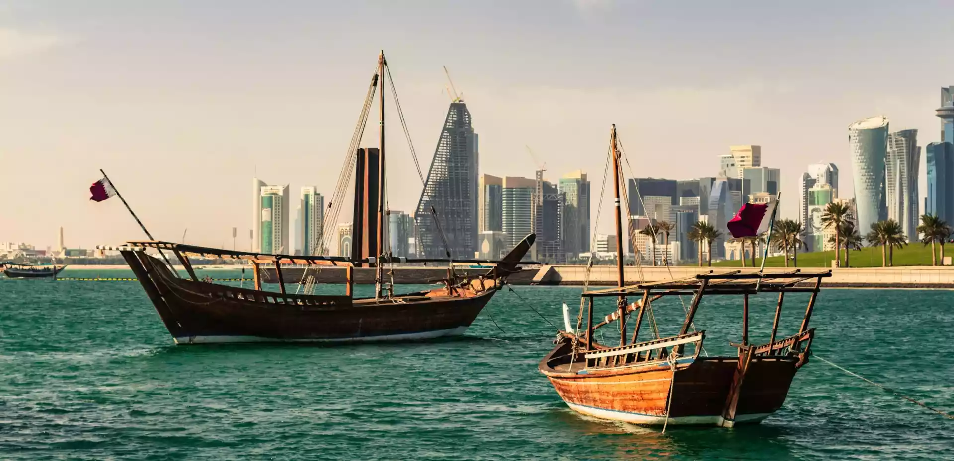 To seilbåter glir stille over vannet ved havnen i Doha, med en klar blå himmel over dem og byens skyline som bakteppe.