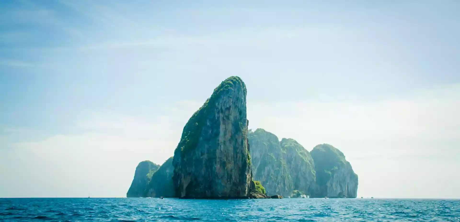 En bratt øy i Thailand strekker seg opp mot en blå himmel fra det klare, blå havet.