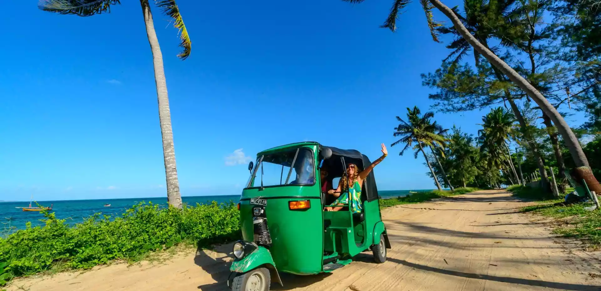 En smaragdgrønn tuk-tuk snor seg langs en sandete vei, omringet av svaiende palmer med det azurblå havet og himmelen som danner en fortryllende bakgrunn.