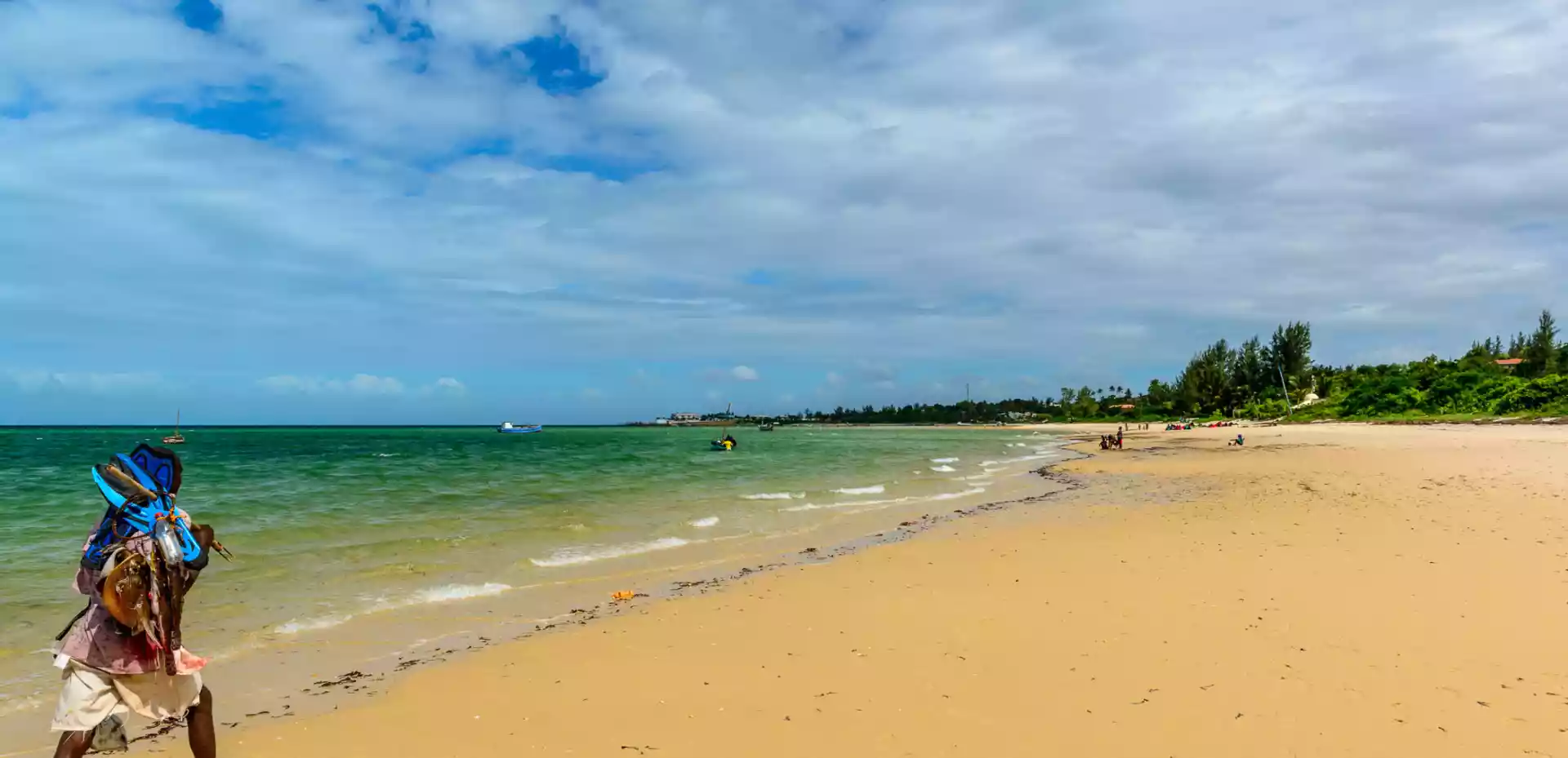 En mann vandrer langs kysten av Mosambik, med det dype blå havet som bakteppe og den myke sandstranden som strekker seg foran ham, en harmonisk scene av ro og skjønnhet.