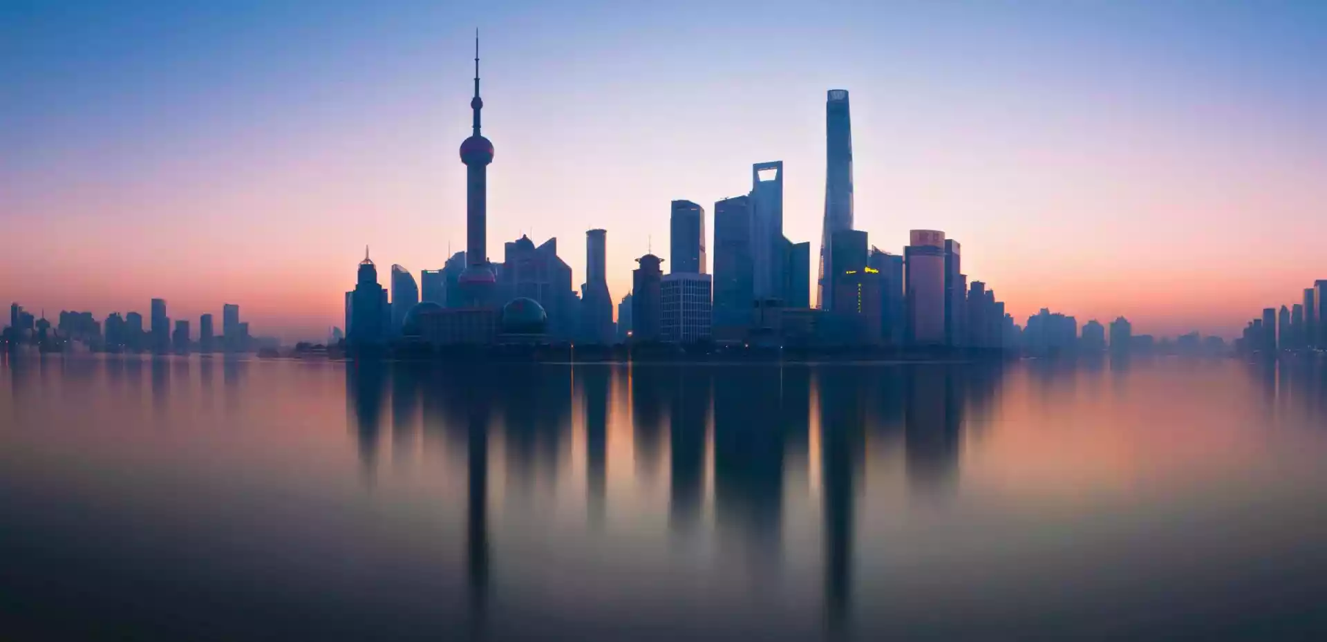 Bilde av skylinen i Shanghai tatt fra vannet. I bakgrunnen blir lyst opp av en oransje solnedgang, vannet og skyskraperne er mørkt.