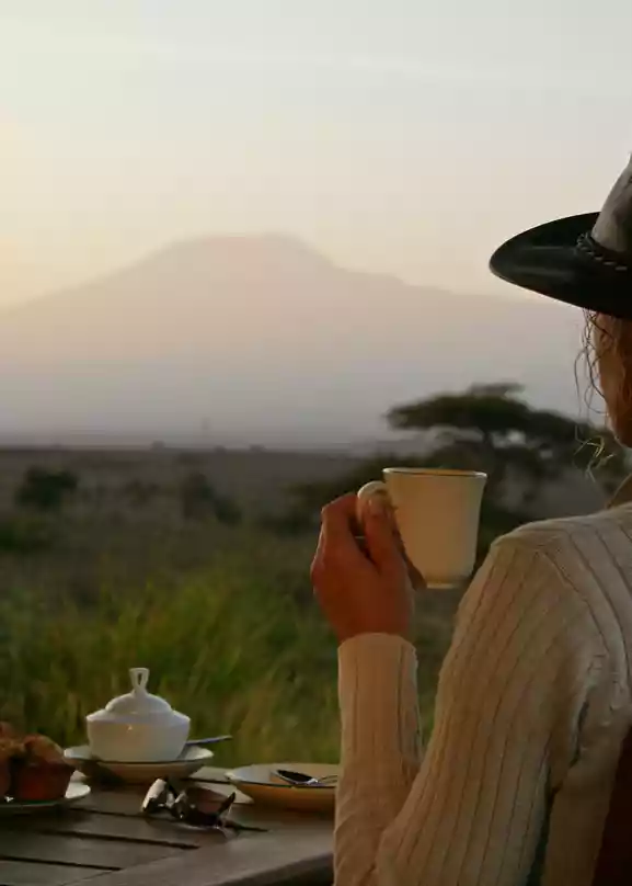Dame i lærhatt drikker kaffi og sitter ved et trebord med scones og en lattern, mens den afrikanske savannen skinner i bakgrunnen.