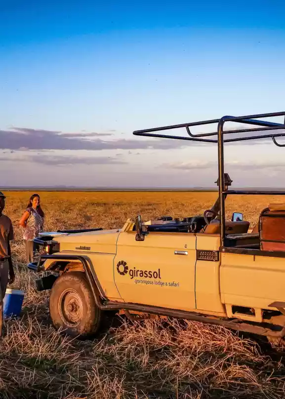 To smilende personer står ved siden av en safari jeep på den vidstrakte savannen i Gorongosa, Mosambik, med en klar blå himmel som bakgrunn. Deres ansiktsuttrykk utstråler glede og spenning mens de utforsker dette naturskjønne området. Det er et øyeblikk av ren lykke og frihet fanget i dette bilde, som minner oss om gleden ved å oppdage det ukjente og dele eventyret med noen vi er glad i.