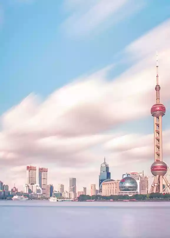 Bilde av skylinen i Shanghai tatt fra vannet. Rosa skyer på en blå himmel over høye skyskrapere over et blått vann.