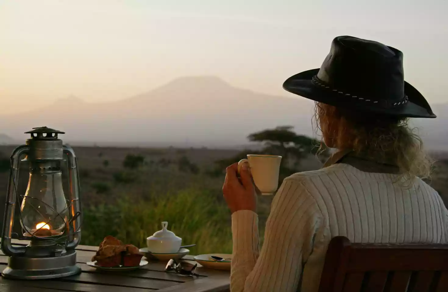 Dame i lærhatt drikker kaffi og sitter ved et trebord med scones og en lattern, mens den afrikanske savannen skinner i bakgrunnen.