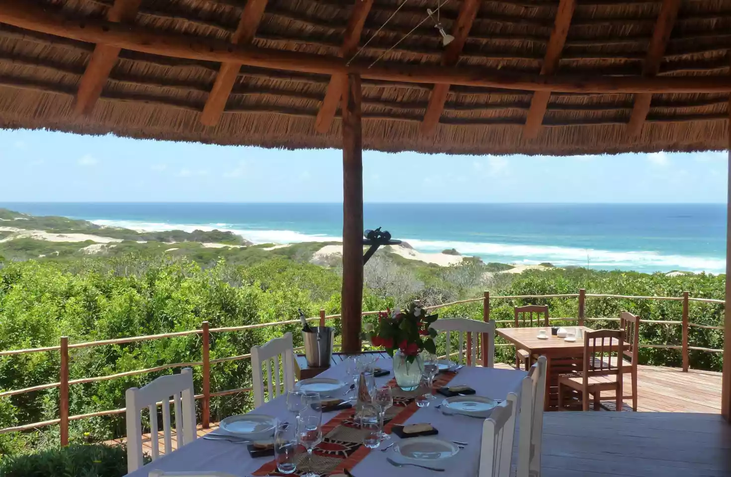 Et elegant dekket bord med hvit duk på Dunes de Dovela Eco Lodge. Stråtaket gir skygge over bordet, mens den spektakulære utsikten viser havet, frodig natur og en klar blå himmel i bakgrunnen.