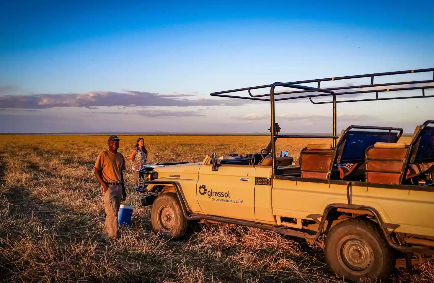 To smilende personer står ved siden av en safari jeep på den vidstrakte savannen i Gorongosa, Mosambik, med en klar blå himmel som bakgrunn. Deres ansiktsuttrykk utstråler glede og spenning mens de utforsker dette naturskjønne området. Det er et øyeblikk av ren lykke og frihet fanget i dette bilde, som minner oss om gleden ved å oppdage det ukjente og dele eventyret med noen vi er glad i.