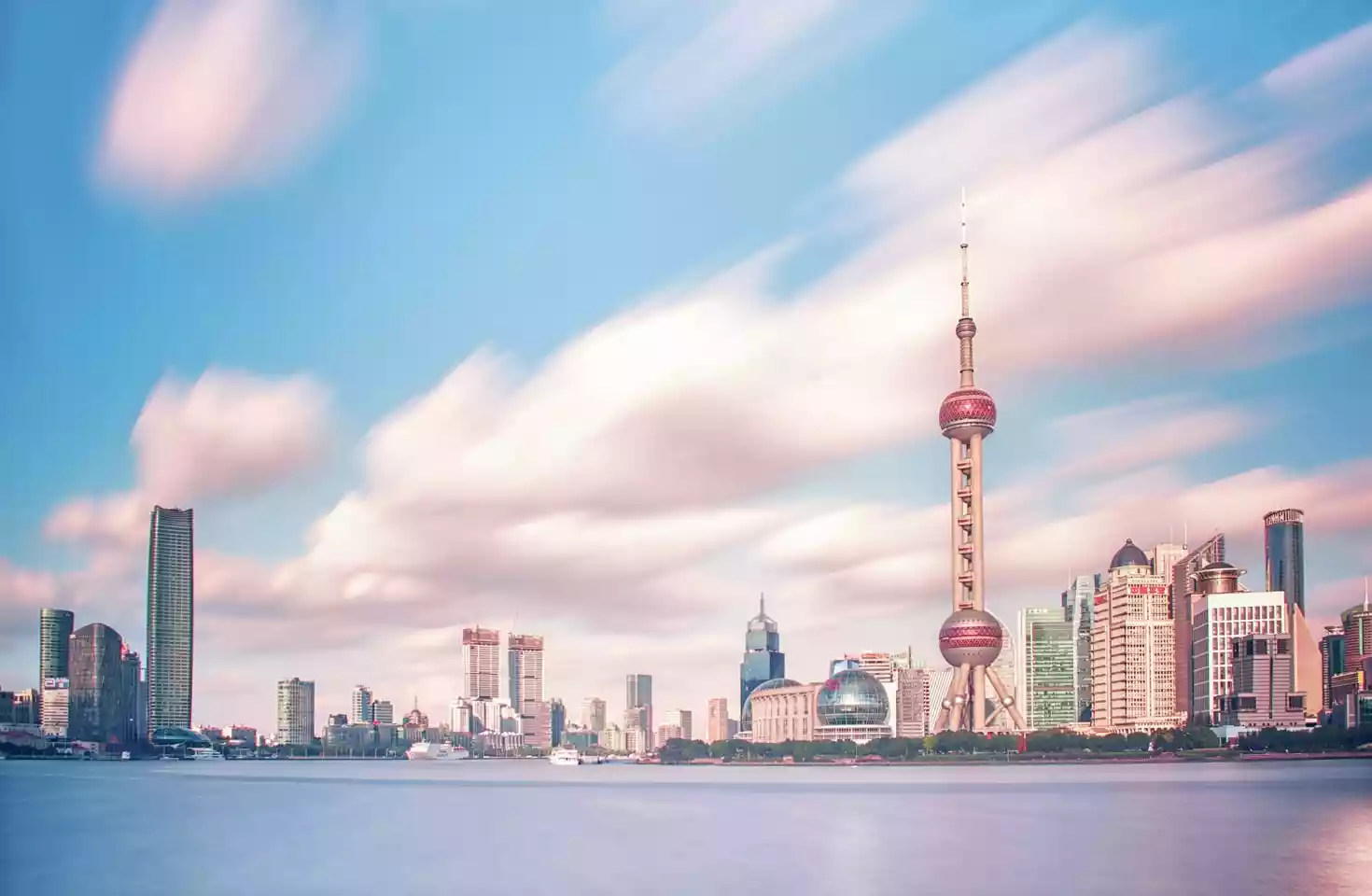 Bilde av skylinen i Shanghai tatt fra vannet. Rosa skyer på en blå himmel over høye skyskrapere over et blått vann.