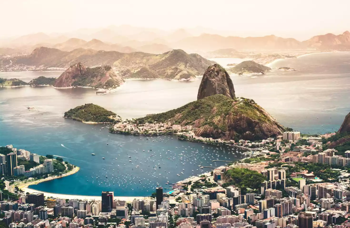 Rio de Janeiro i solnedgangen. Høye frodige klipper, blågrått hav dekket av små båter langs havnen, og Rio de Janairo i forgrunnen med høye sement blokker.