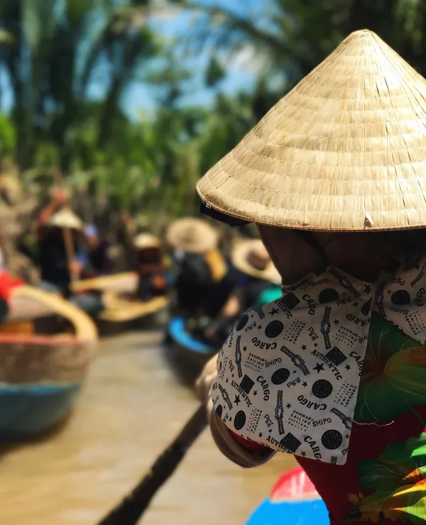 En person ror en kajakk på Mekong elven, med flere kajakker i bakgrunnen. De fleste personene i kajakkene har på seg en tradisjonell vietnamesisk strå hatt.