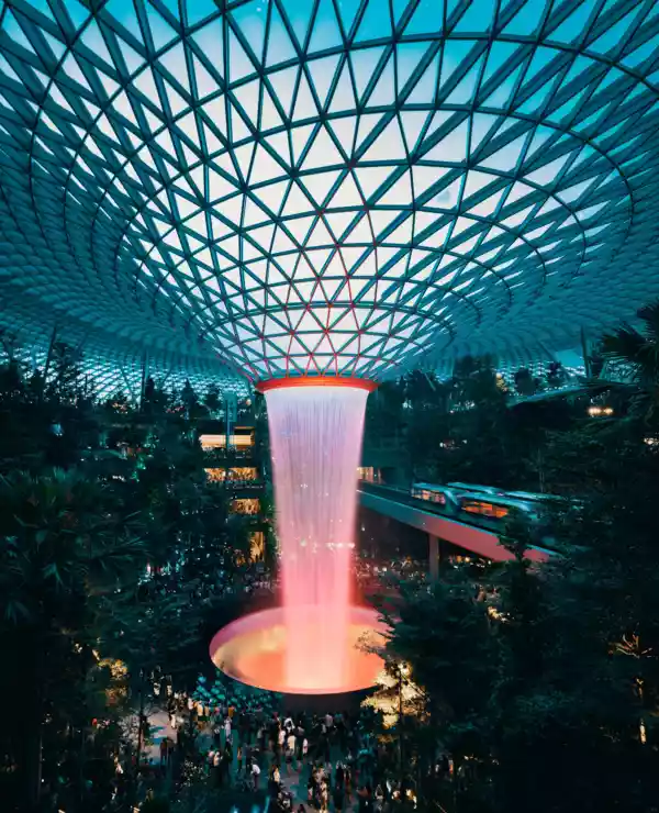 Innsiden av Singapore lufthavn. Taket er formet som en opp ned kjegle, hvor et oransje fossefall renner ned fra toppen av kjeglen. Rommet er fylt av grønn vegetasjon.