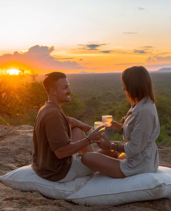 En man og en dame, sitter på hver sin pute og skåler med noe godt å drikke. De er omgitt av to latterner, vakker afrikansk natur og en solnedgang.