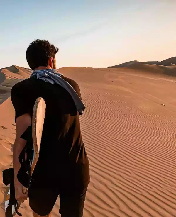 En man i en ørken som bærer et surfebrett, mens han ser til høyre.