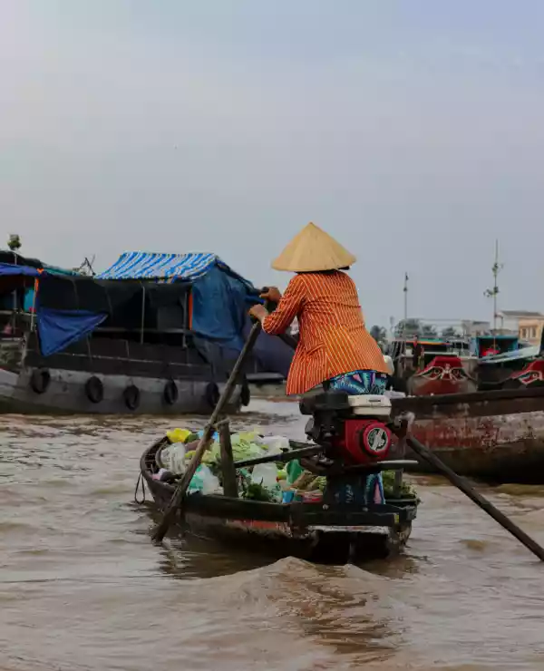 En person i en tradisjonell vietnamesisk strå hatt står i en liten båt på vei til et flyttende marked.
