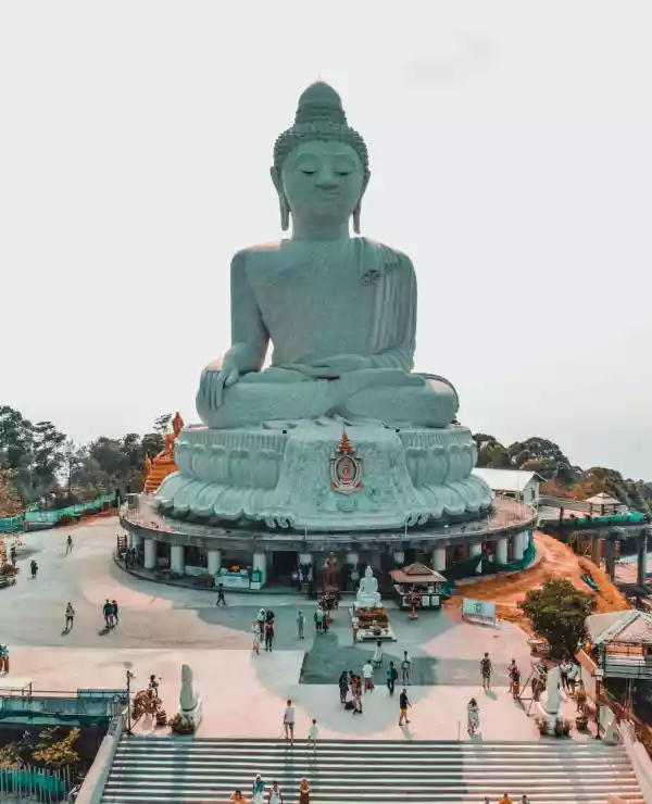 En Buddhastatue på et platå i Phuket, omgitt av mange mennesker, med en klar himmel i bakgrunnen.