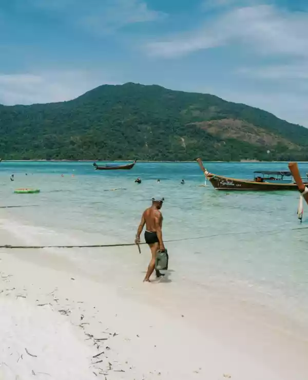 En mann nyter strandlivet på en thailandsk strand, hvor båter ligger fortøyd langs kysten og et frodig fjell strekker seg i bakgrunnen. Den hvite sanden strekker seg foran ham, mens den klare blå himmelen skaper en perfekt ramme rundt scenen.