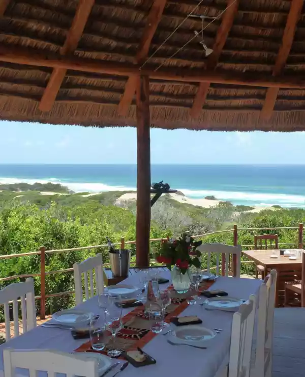 Et elegant dekket bord med hvit duk på Dunes de Dovela Eco Lodge. Stråtaket gir skygge over bordet, mens den spektakulære utsikten viser havet, frodig natur og en klar blå himmel i bakgrunnen.