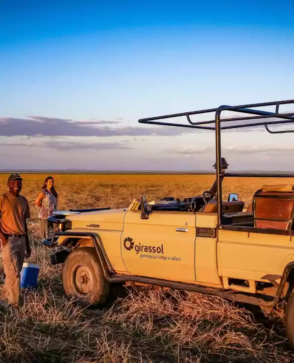 To smilende personer står ved siden av en safari jeep på den vidstrakte savannen i Gorongosa, Mosambik, med en klar blå himmel som bakgrunn. Deres ansiktsuttrykk utstråler glede og spenning mens de utforsker dette naturskjønne området. Det er et øyeblikk av ren lykke og frihet fanget i dette bilde, som minner oss om gleden ved å oppdage det ukjente og dele eventyret med noen vi er glad i.