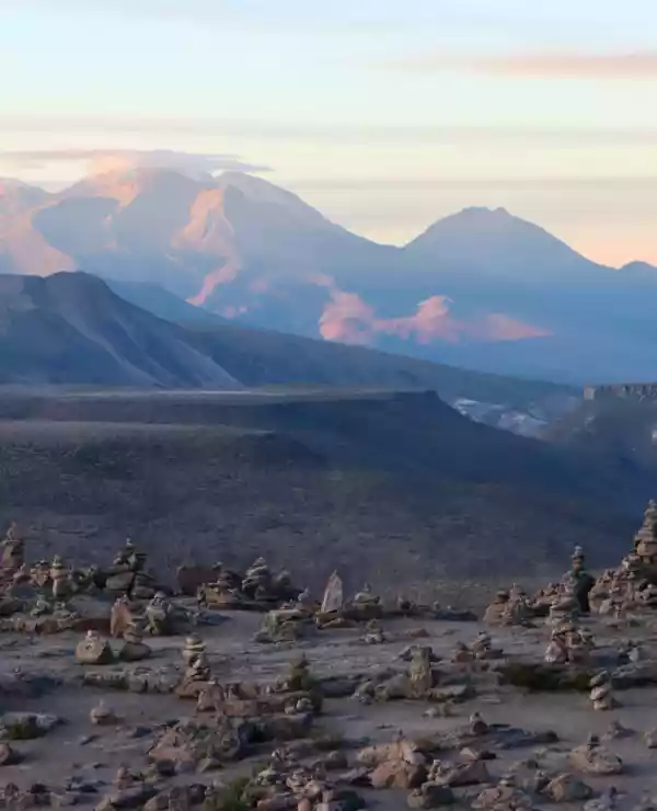 Panoramautsikt over det bølgende landskapet i Colca Canyon, med snødekte fjell i bakgrunnen. En blå himmel prydet med oransje skyer kompletterer den imponerende naturen.