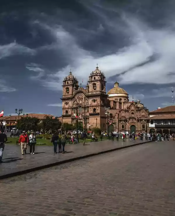 Barokk arkitektur i Cusco med brolagt gate i forgrunnen og mennesker som går over gangfeltet under en klar blå himmel.