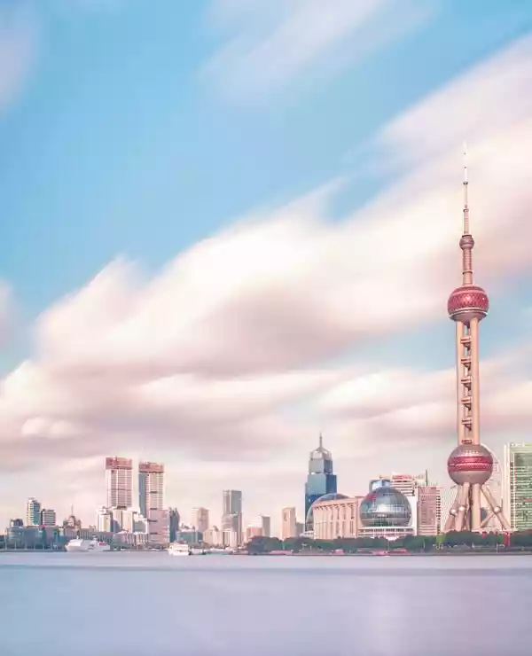 Bilde av skylinen i Shanghai tatt fra vannet. Rosa skyer på en blå himmel over høye skyskrapere over et blått vann.