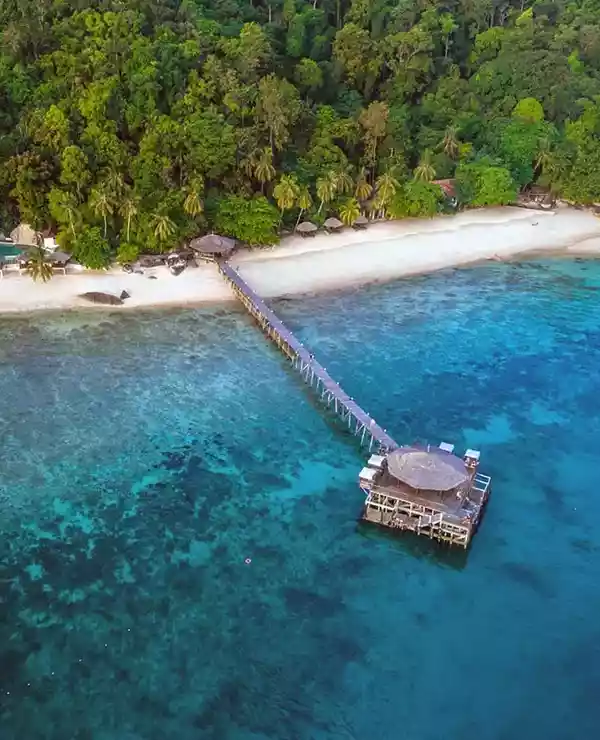 Overblikk over Japamala Resort. En bygning på et blått hav med en gangbru til en strandlinje og frodig skog, med tre bygninger i skogen.